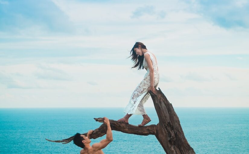 woman sitting on tree trunk with man holding on branch near sea under white clouds during daytime